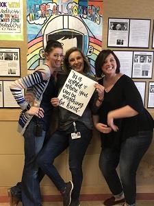 3 teachers posed in front of a bulletin board holding a sign about being kind.